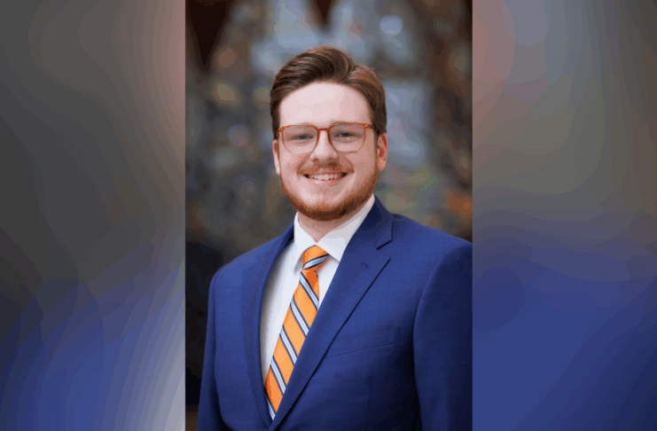 A man with glasses and a beard smiles, wearing a blue suit, white shirt, and orange-striped tie, against a blurred background.