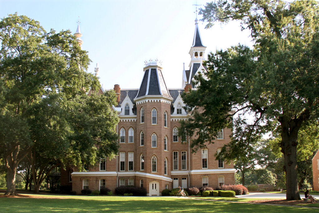A historic, three-story brick building with steep towers and white trim sits surrounded by large trees and green lawn.