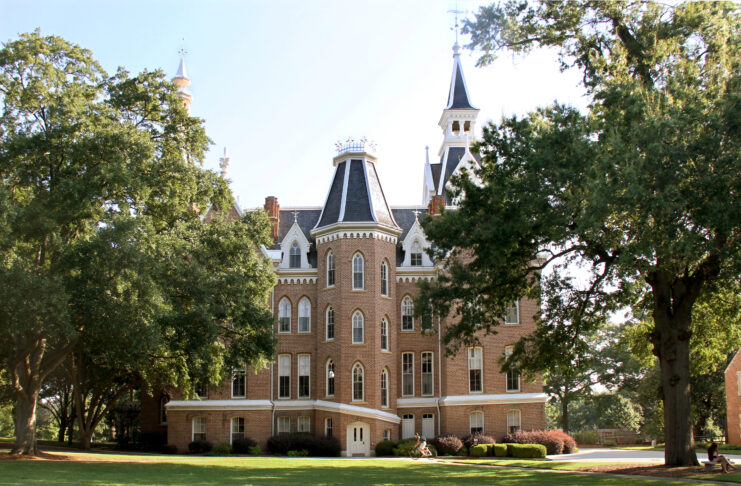 A historic, three-story brick building with steep towers and white trim sits surrounded by large trees and green lawn.