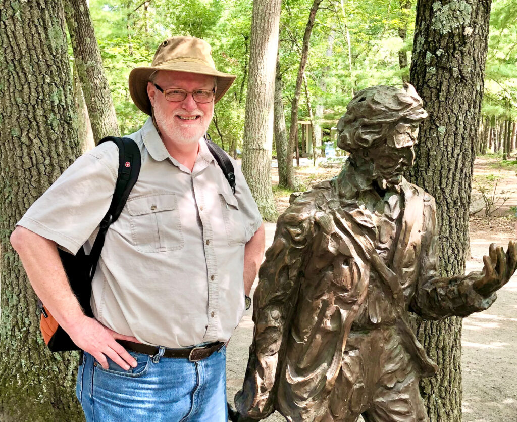 Dr. Marc Jolley stands next to a bronze statue of Henry David Thoreau in a wooded outdoor setting.