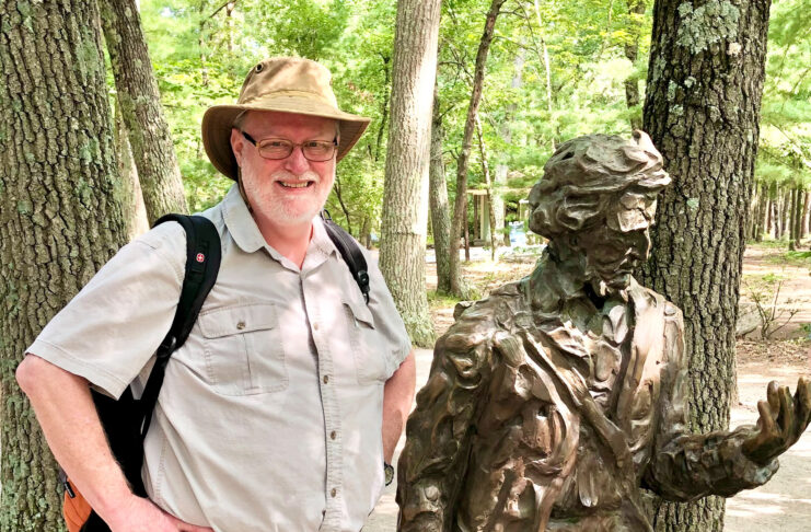 Dr. Marc Jolley stands next to a bronze statue of Henry David Thoreau in a wooded outdoor setting.