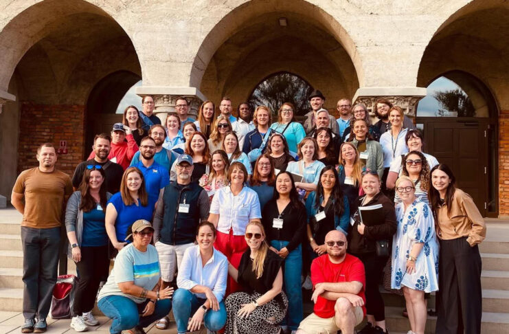 A large group of adults poses together on outdoor steps in front of an arched stone building.