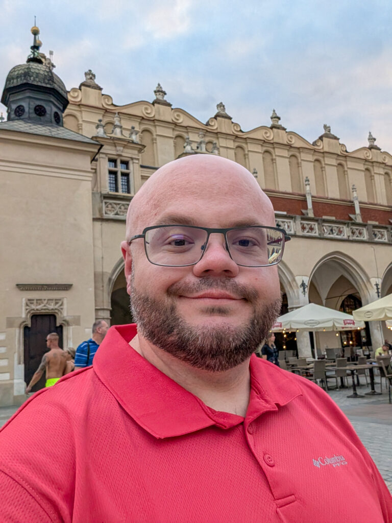 A man in a red shirt and glasses stands in front of a historic building with arches and outdoor seating.