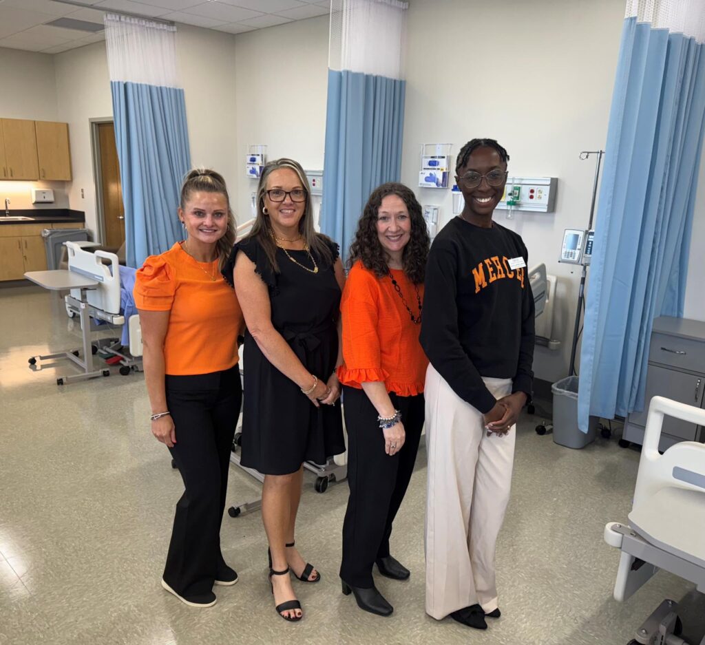 Four women stand in a clinical room, smiling at the camera, with hospital beds and medical equipment in the background.