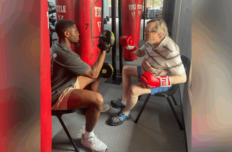 An older man wearing boxing gloves practices punches with a trainer holding pads in a gym.