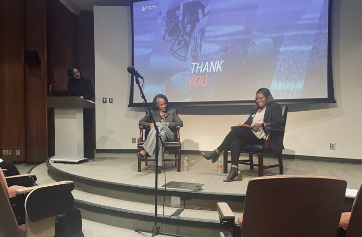 Three women participate in a discussion on stage in an auditorium, with a Thank You slide projected behind them.