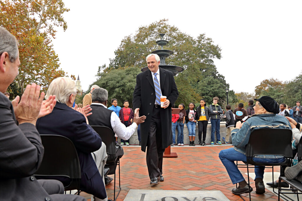Mercer President William D. Underwood walks in front of seated people outdoors, while a group of children stands in the background near a fountain.