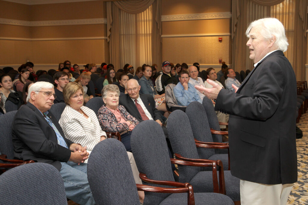 An older man in a suit speaks to an audience seated in rows in a formal, carpeted room.