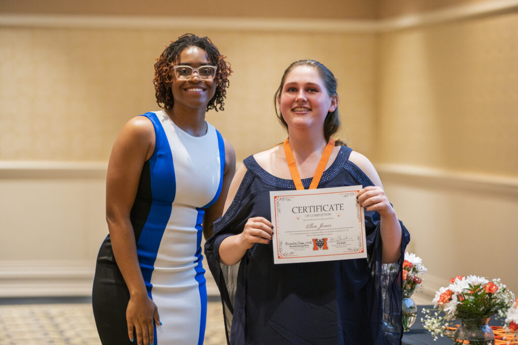 Two women stand indoors; one holds a certificate and wears a medal, both are smiling at the camera.