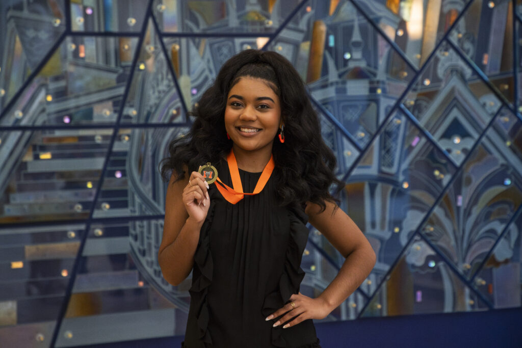 Georgia Taylor smiles while holding a medal on an orange ribbon in front of a geometric background.