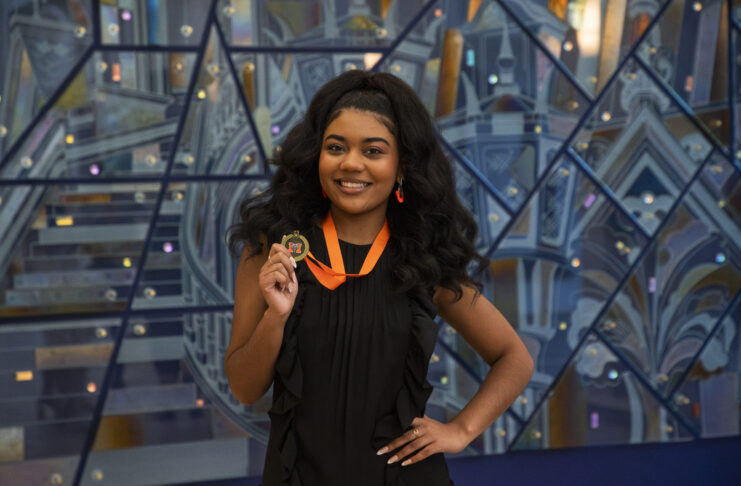 Georgia Taylor smiles while holding a medal on an orange ribbon in front of a geometric background.