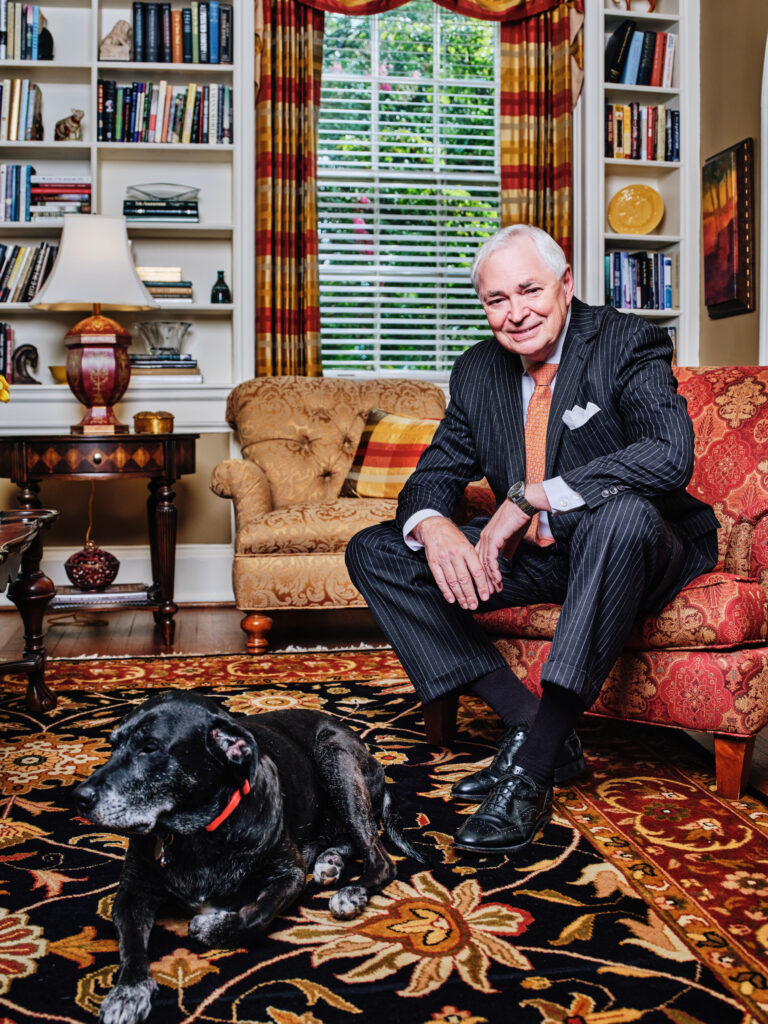 Mercer President William Dr. Underwood sits on a patterned armchair beside a black dog in a well-decorated living room with bookshelves.