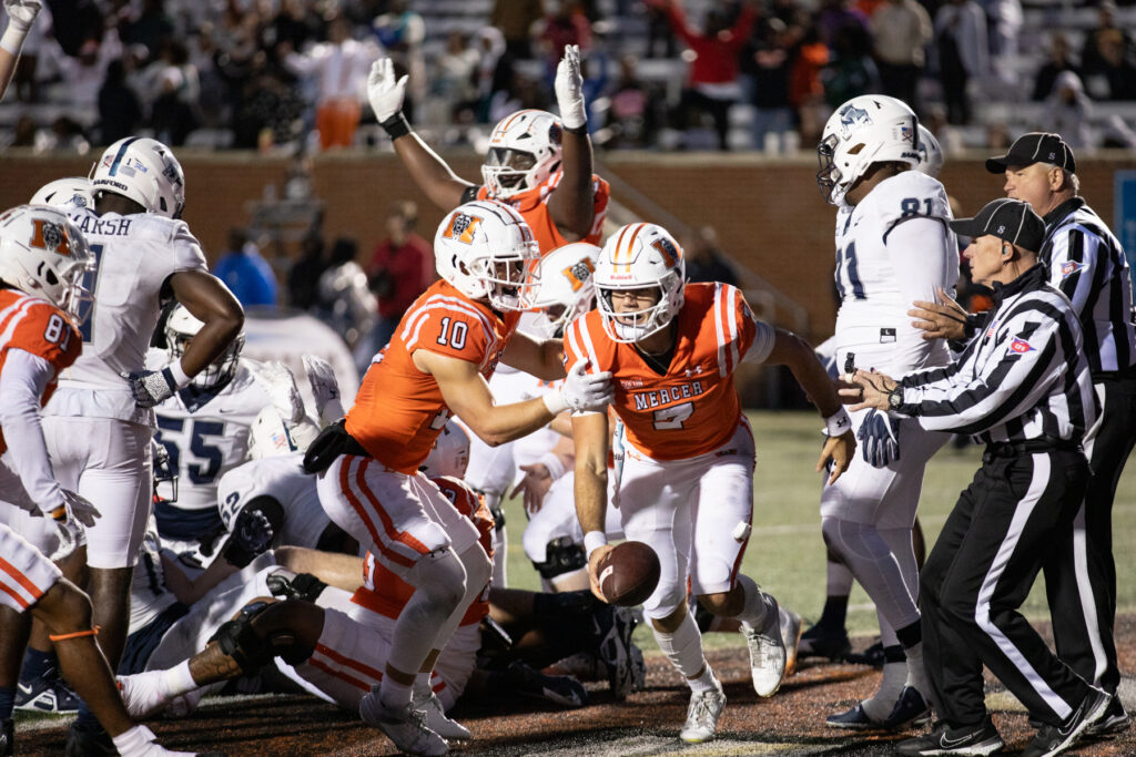 Football players from two teams react as Mercer player in an orange jersey recovers a fumble near the end zone, referees nearby.