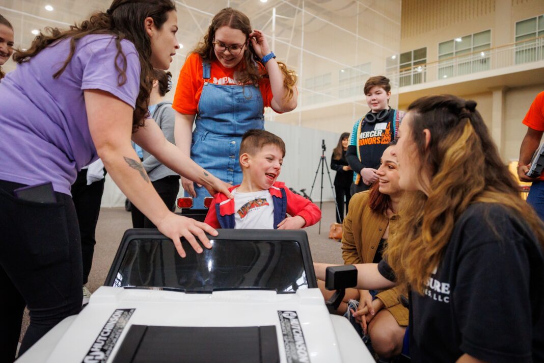 A young boy in a toy car smiles as several adults and children gather around him indoors.