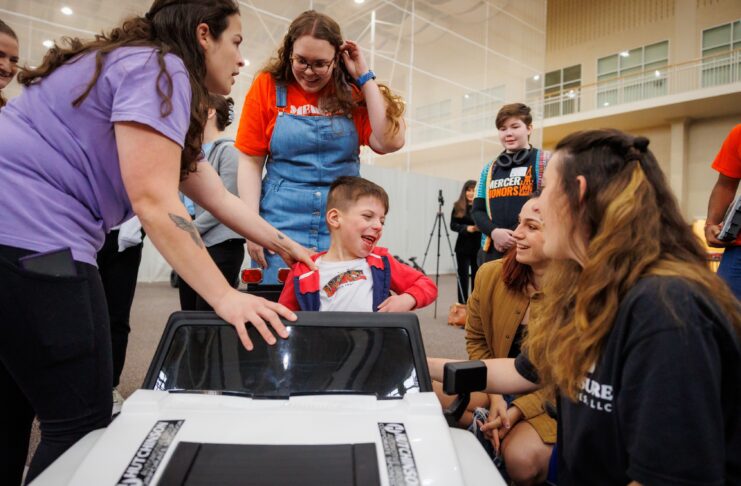 A young boy in a toy car smiles as several adults and children gather around him indoors.
