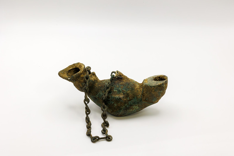 Ancient metal oil lamp with a corroded surface and an attached chain, displayed on a plain white background.