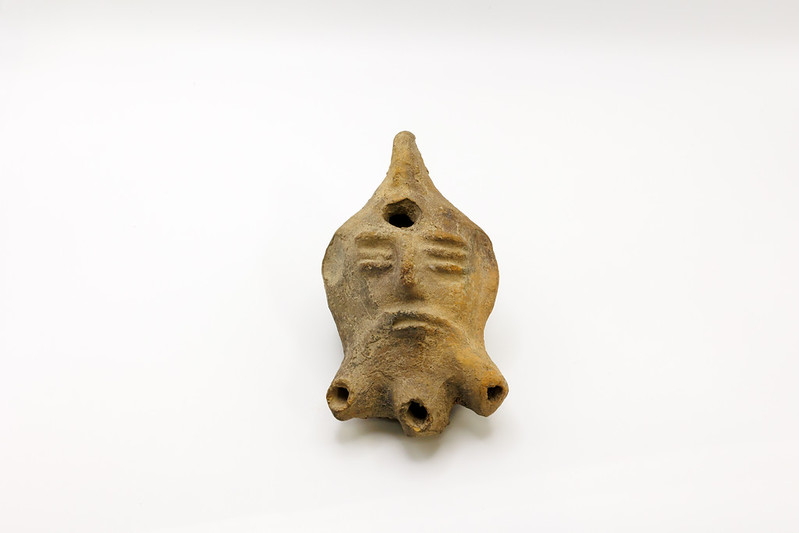 A clay artifact with a face-like design and multiple holes, placed on a plain white background.