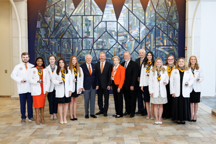 Nathan Deal Scholars 2025 A group of people in white coats and formal attire pose in front of a large stained glass window indoors.
