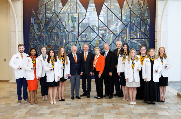 A group of people in white coats and formal attire pose in front of a large stained glass window indoors.