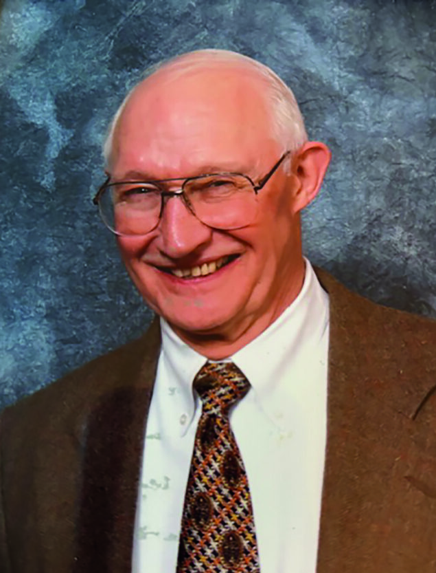 An older man with glasses and a patterned tie smiles, wearing a brown suit jacket, in front of a gray mottled background.