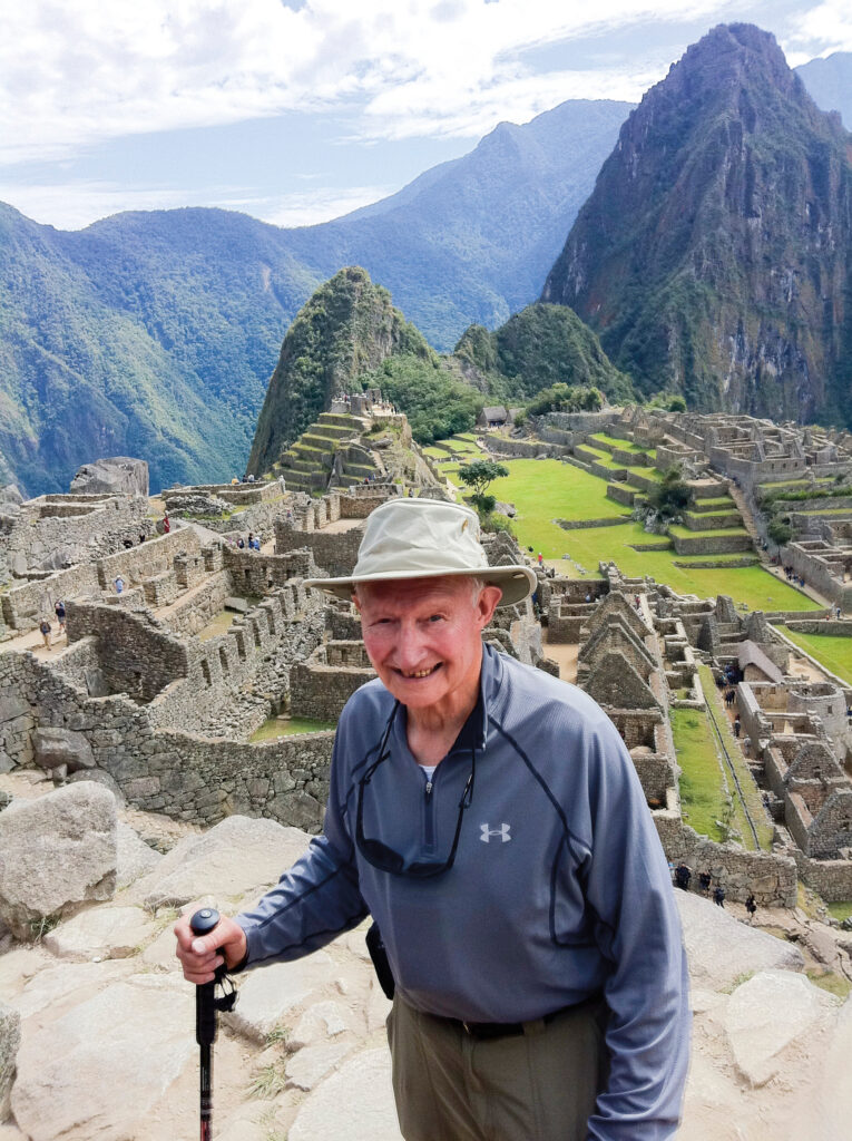 Older man with a walking stick stands in front of ancient ruins at Machu Picchu, surrounded by mountains.