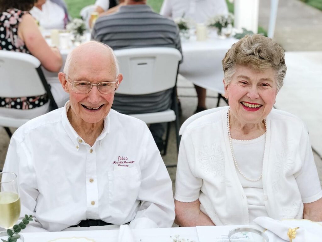 An elderly man and woman sit side by side at an outdoor table, both smiling at the camera.