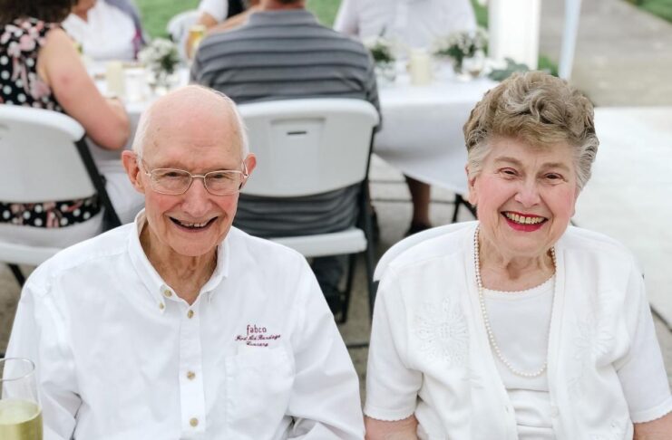 An elderly man and woman sit side by side at an outdoor table, both smiling at the camera.