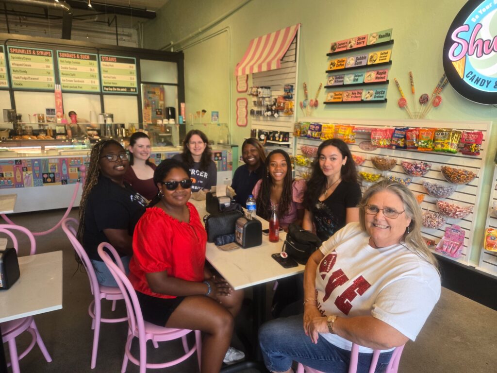 A group of eight women sit around a table at a candy shop, smiling at the camera.