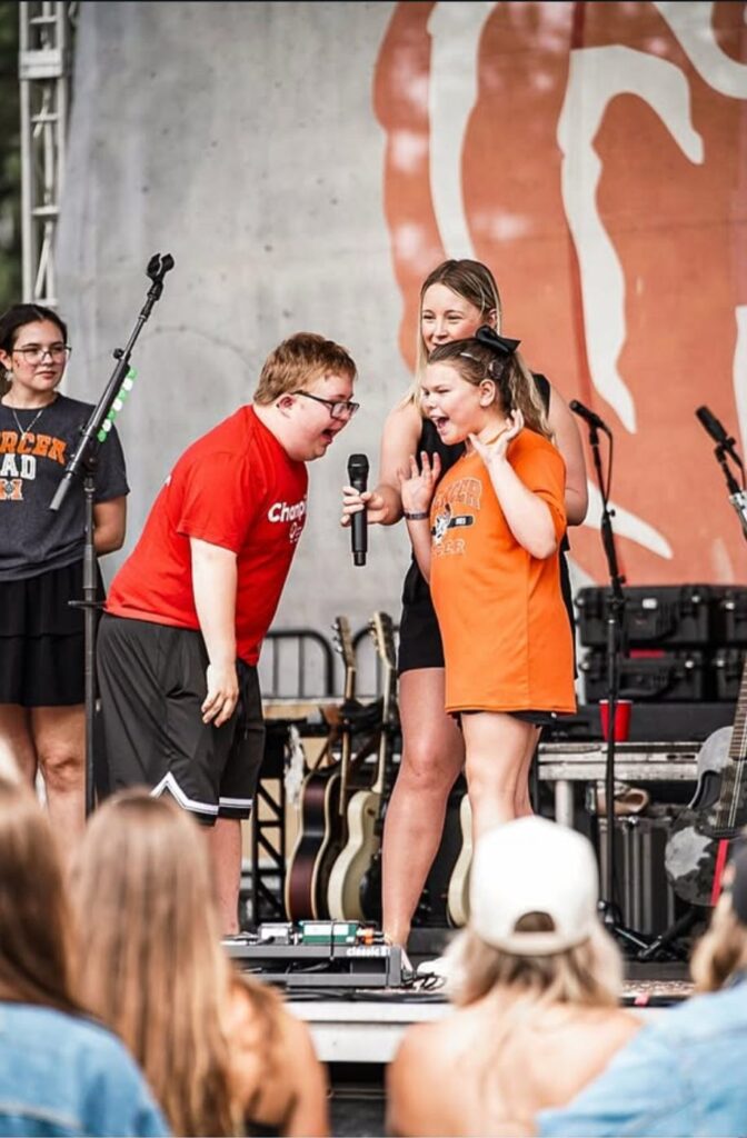Four people stand on an outdoor stage, two holding a microphone, with musical instruments and an audience visible.