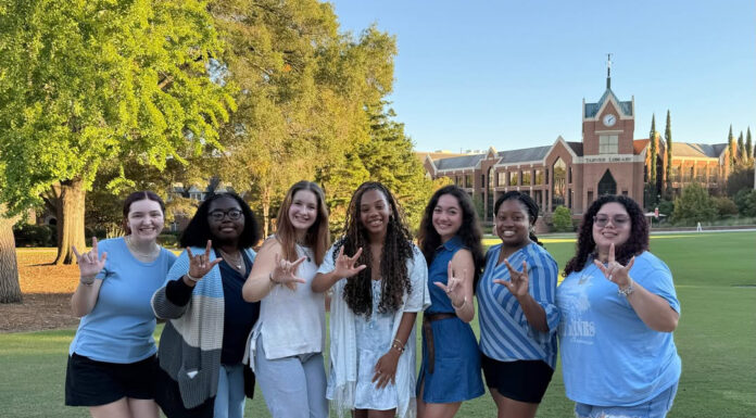 Mercer junior launches club to help students learn American Sign Language Seven people stand outdoors on a path, smiling and making hand signs, with a brick building and trees in the background.