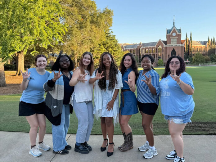 Seven people stand outdoors on a path, smiling and making hand signs, with a brick building and trees in the background.