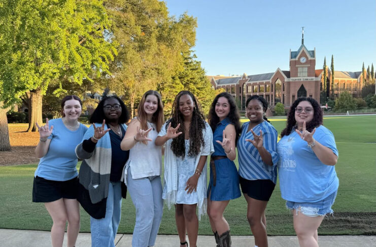 Seven people stand outdoors on a path, smiling and making hand signs, with a brick building and trees in the background.