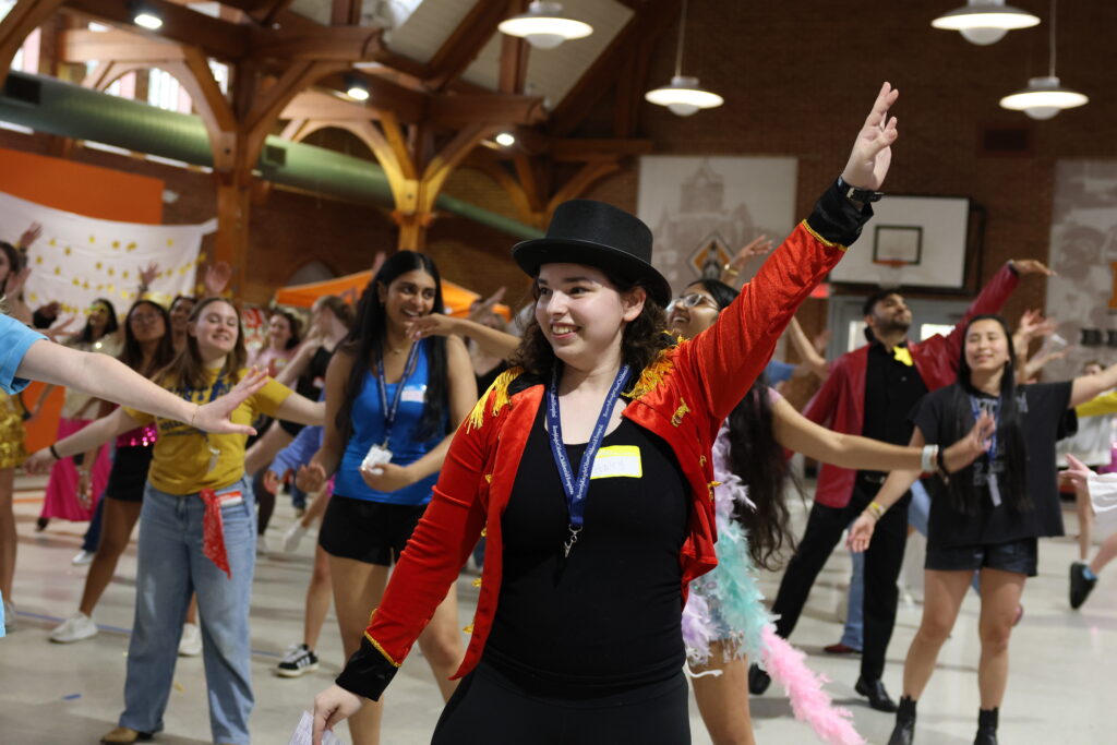 A group of people dance indoors as a person in a red jacket and top hat leads with an arm raised.