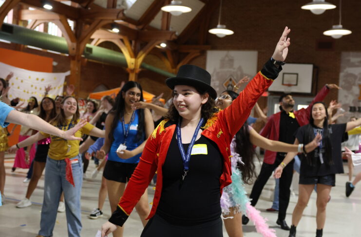 A group of people dance indoors as a person in a red jacket and top hat leads with an arm raised.