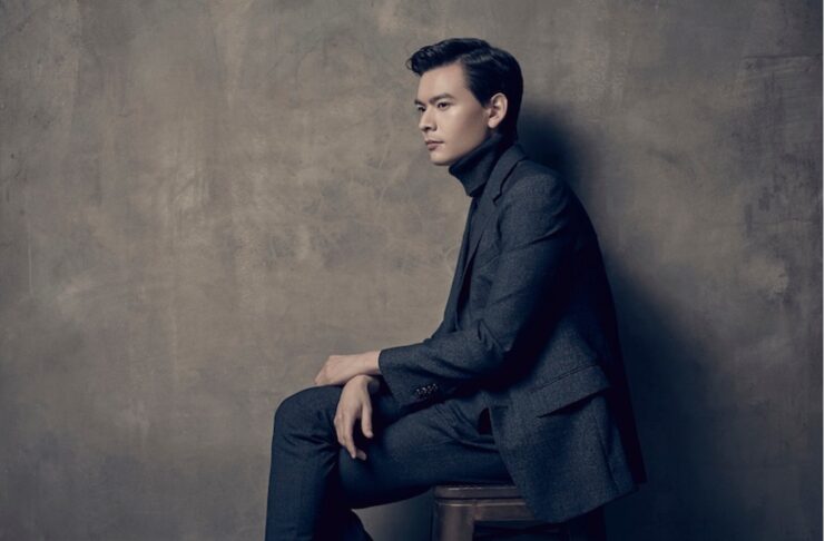 A man in a dark suit sits sideways on a stool against a textured gray wall, looking ahead with a neutral expression.
