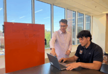 Biology professor earns EXPy Award for gene project work Two people work together at a table; one points at a laptop while the other types, with a whiteboard in the background.
