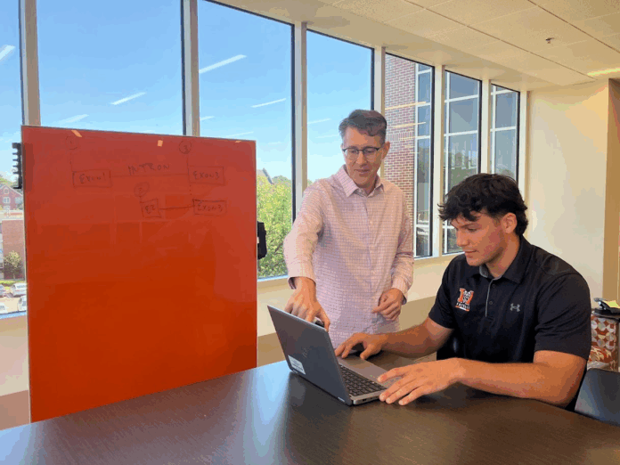 John Stanga Two people work together at a table; one points at a laptop while the other types, with a whiteboard in the background.