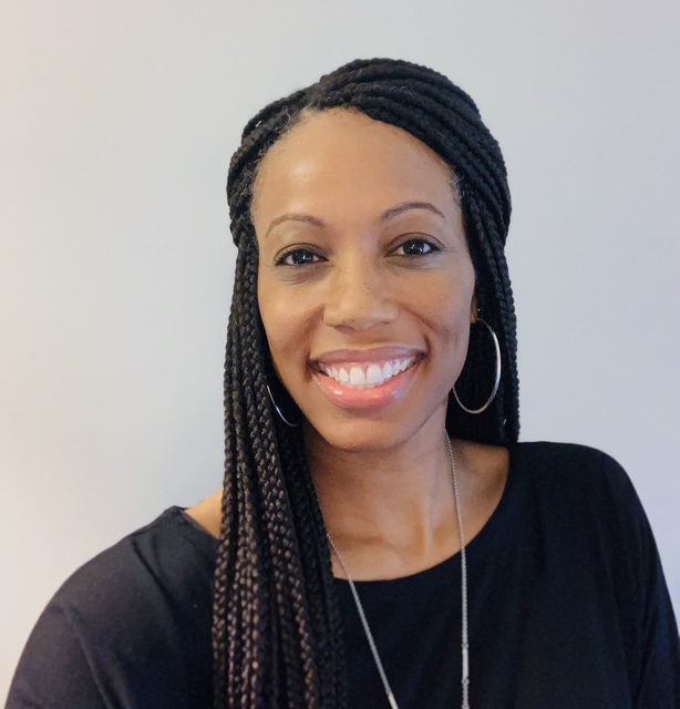 A woman with long braided hair, wearing hoop earrings and a black top, smiles at the camera against a plain background.