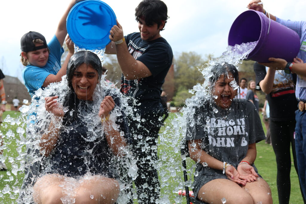 Two people sitting outdoors react as others pour buckets of water over them during a group activity on grass.