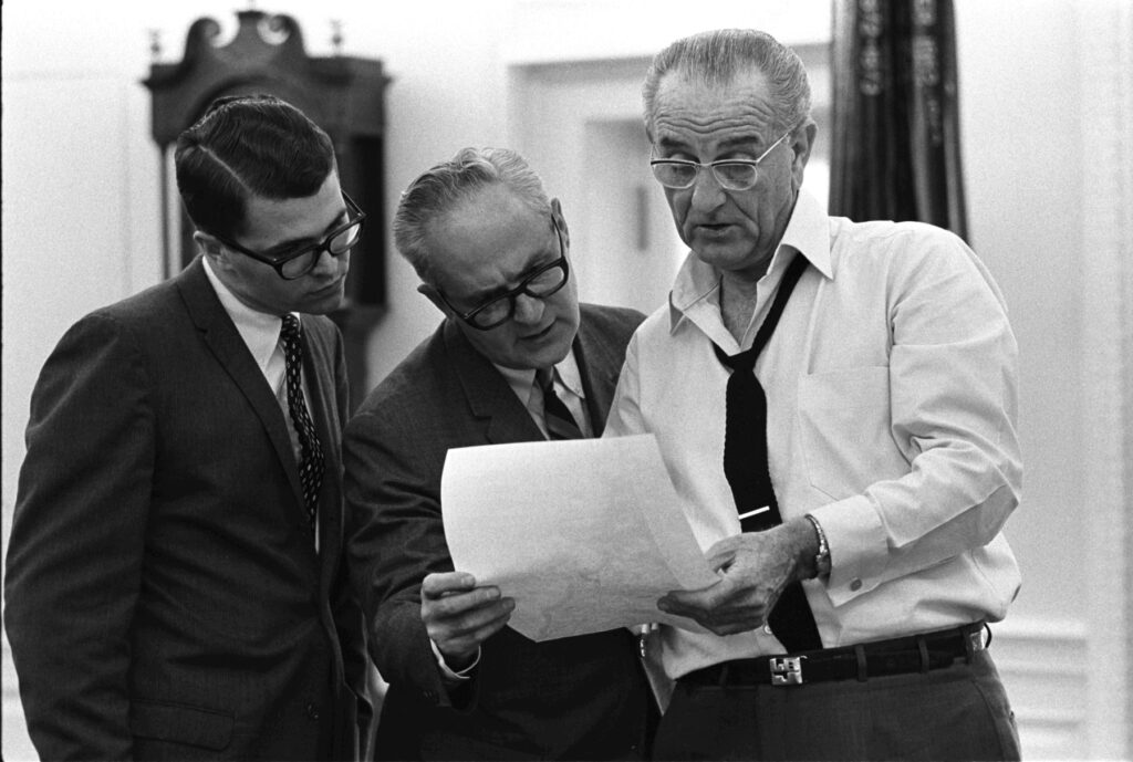 Three men in suits examine a document together in an office setting.
