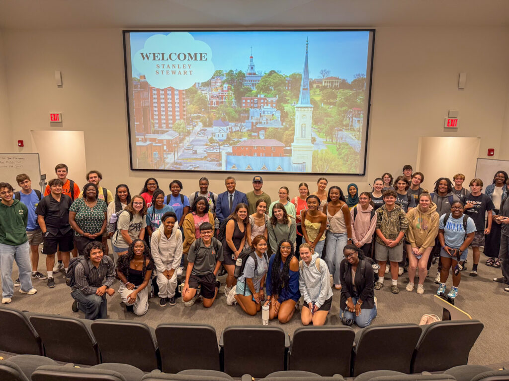 A large group of students poses together in an auditorium with a welcome message projected on the screen behind them.