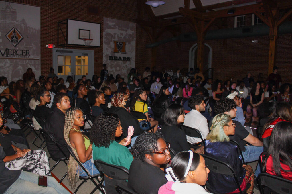 A large group of people sit in rows of chairs facing forward at an indoor event, with a Mercer Bears banner on the wall.