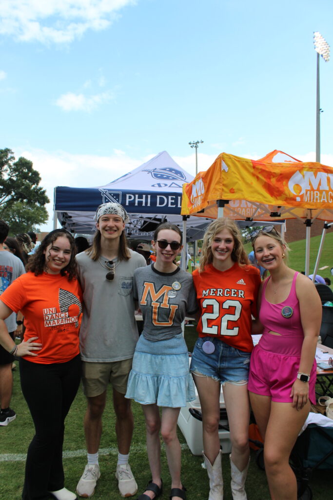 Five young adults stand together smiling in front of event tents outdoors on a sunny day.