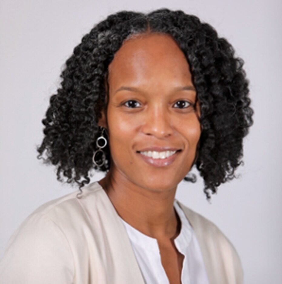 Woman with curly hair wearing a light top and beige cardigan, smiling at the camera against a plain background.