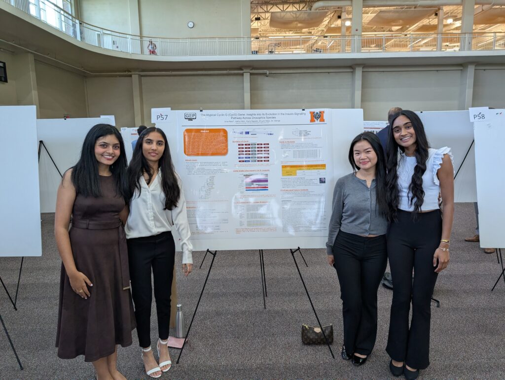 Four women stand in front of a scientific research poster displayed on an easel at a poster presentation event.