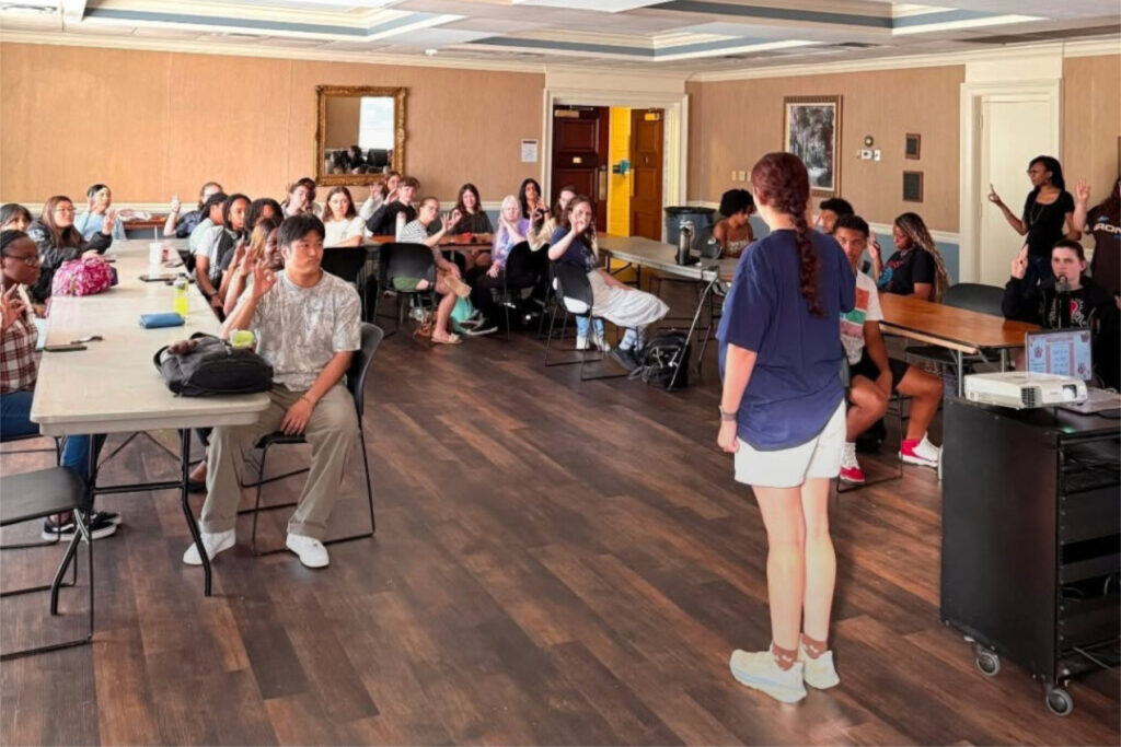 A woman stands speaking to a seated group of people in a conference room with tables and a projector.