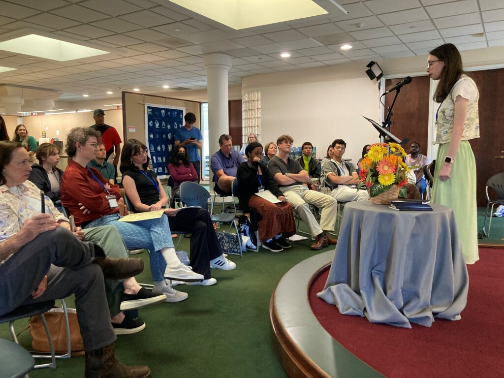 A woman stands at a podium speaking to an audience seated in a room with a flower arrangement on a small table.
