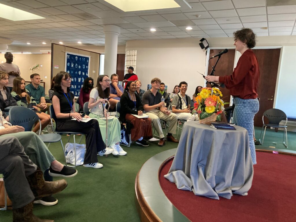 A person speaks at a podium to an audience seated in a room, with a table of flowers in the foreground.