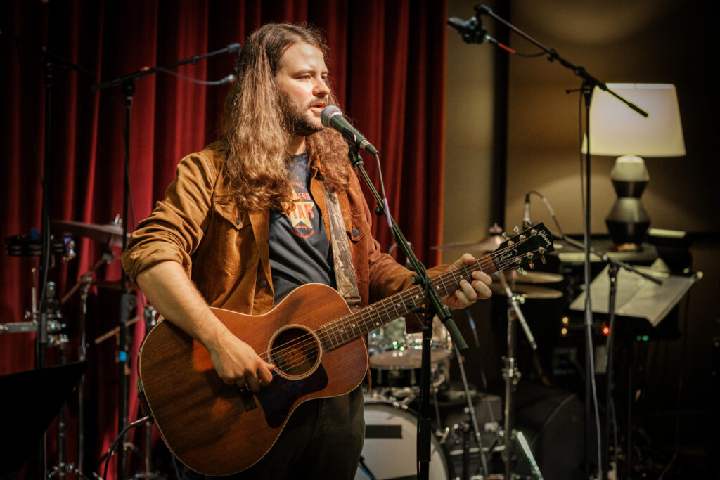 Brent Cobb plays an acoustic guitar and sings into a microphone on a stage with red curtains and musical equipment.