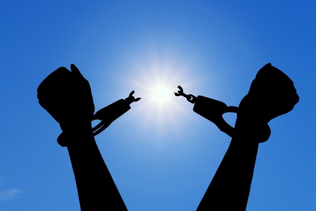 Silhouette of hands breaking free from handcuffs against a bright sun in a clear blue sky.